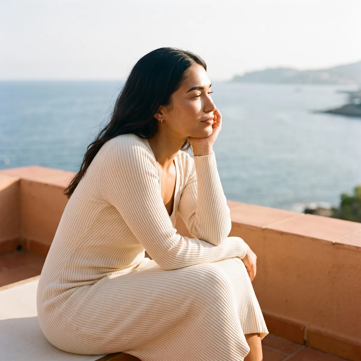 A woman in a cream knit dress on a sunlit balcony, her cheek resting on her hand, skin firm and luminous