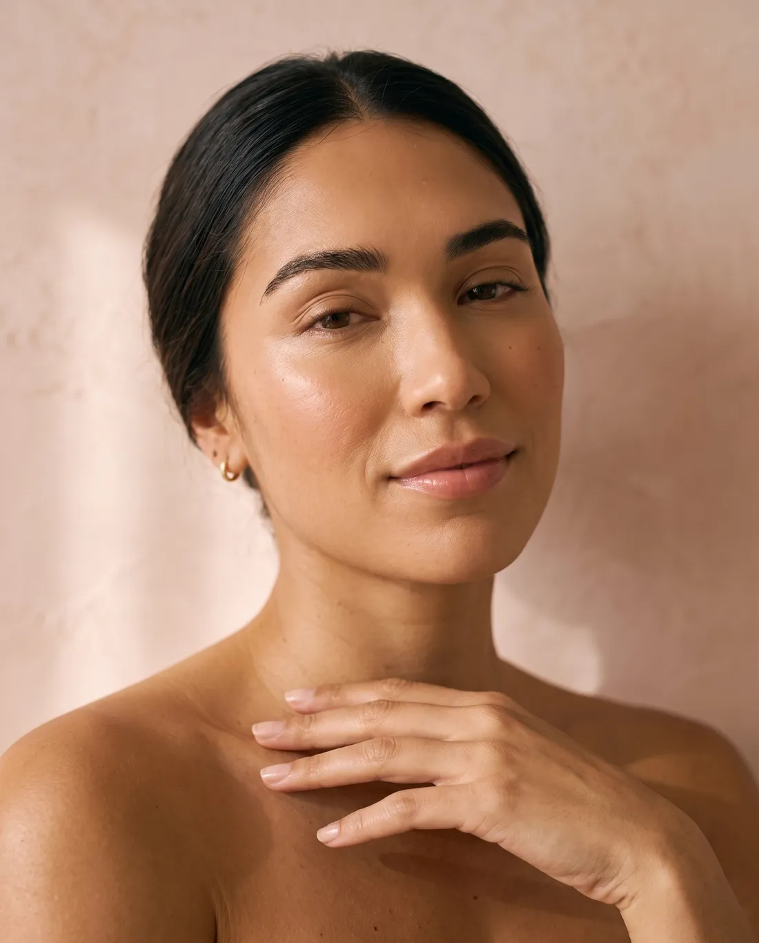 A woman against a soft blush wall, fingertips at her collarbone, her face refreshed and radiant